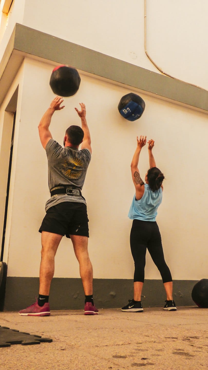 Two people perform wall ball exercises against a building wall.
